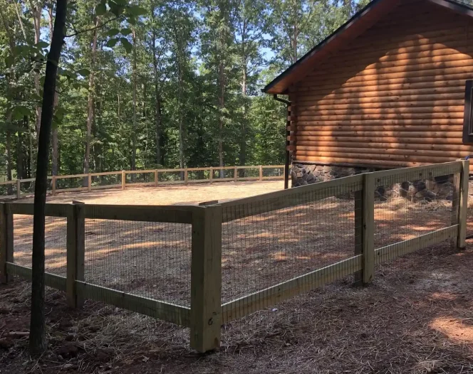 A newly built wooden fence with wire mesh enclosing a sandy area next to a log cabin, surrounded by trees.